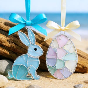 May include: Two Easter ornaments: a blue glass bunny and a stained-glass egg. The bunny is made of wire and blue glass, with a blue ribbon. The egg is made of wire and pastel-colored glass, with a yellow ribbon. Both are hanging.