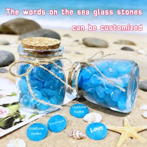 Puede incluir: Dos frascos de vidrio llenos de piedras de vidrio de mar azules, uno en posición vertical y otro de lado, atados con hilo. Los frascos están sobre una superficie arenosa con piedras azules y conchas marinas. El texto en las piedras dice "custom name", "Veronique" y "Love". El texto "The words on the sea glass stones can be customized" también es visible.