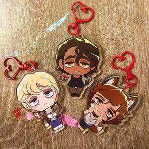 May include: Three acrylic keychains featuring cartoon characters with heart-shaped clasps. The characters have various hair colors and outfits. The keychains are on a wooden surface with paint splatters.