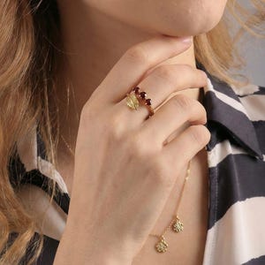 Pode incluir: Um anel de ouro com um design de folha e três pedras preciosas vermelhas. Um colar de ouro com dois pingentes de flores também é visível. A pessoa está usando uma camisa listrada em preto e branco.