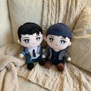 Könnte beinhalten: Zwei Plüschpuppen von Charakteren im Anime-Stil. Eine Puppe trägt eine blaue Jacke, ein weißes Hemd und eine schwarze Krawatte. Die andere Puppe trägt eine graue Jacke und eine blaue Mütze. Beide Puppen haben schwarzes Haar und hellblaue Augen.