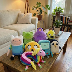 May include: A selection of Adventure Time character plush toys, including Finn, Jake, and BMO, displayed on a wooden coffee table. The toys are brightly coloured and set against a living room backdrop, with a sofa and plants visible.