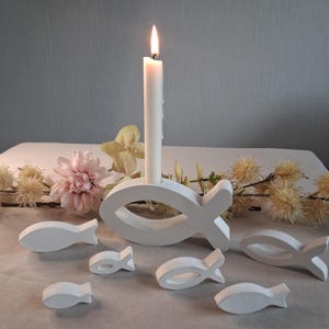 Peut inclure: Une bougie blanche allumée dans un porte-bougie en forme de poisson, entourée de petits poissons blancs. L'ensemble est posé sur une surface blanche, avec des décorations florales, créant une ambiance sereine et décorative.