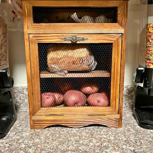 May include: A wooden bread box with a mesh front, holding bread and potatoes. The box has a decorative handle and is filled with various food items. A package of English muffins sits on top.