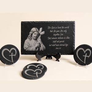Puede incluir: Una placa rectangular de pizarra negra con una imagen grabada de Jesús sosteniendo una cruz y el texto John 3:16, For God so loved the world that He gave His only begotten Son, That whoever believes in Him shall not perish but have eternal life. Tres posavasos redondos de pizarra con diseños grabados de corazones y cruces están dispuestos frente a la placa.