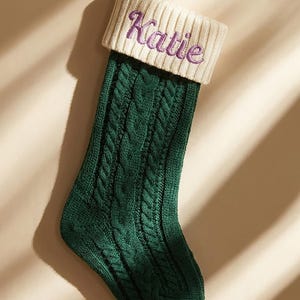 May include: A green cable-knit Christmas stocking with a cream-coloured cuff. The name "Katie" is embroidered in purple script on the cuff. A loop is attached for hanging. A festive holiday decoration.