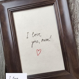 Puede incluir: Una obra de arte enmarcada con el mensaje escrito a mano "I love you, mom!" en tinta negra, acompañado de un pequeño corazón rojo. El marco es de color marrón oscuro y la obra se muestra sobre un lienzo crema.
