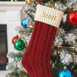 May include: A red cable-knit Christmas stocking with a cream-colored cuff embroidered with the name "Scott" in gold. The stocking hangs on a decorated Christmas tree with colorful ornaments.