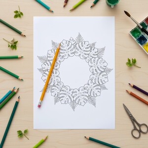 Op de afbeelding: Een witte kleurplaat met een cirkelvormig ontwerp van monstera bladeren en varens. Een gele potlood ligt op de pagina, omringd door kleurpotloden, verf, een penseel en een schaar. De sc&egrave;ne speelt zich af op een licht houten oppervlak.