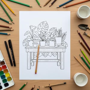 Op de afbeelding: Een zwart-wit lijntekening van potplanten op een houten tafel, klaar om in te kleuren. De tekening is omgeven door kleurpotloden, een aquarelpalet, twee kleine witte kopjes en tuingereedschap op een lichtbruin houten oppervlak.