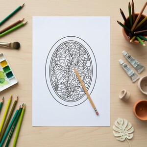 Op de afbeelding: Een zwart-wit lijntekening van een botanische sc&egrave;ne, met verschillende planten in een ovale lijst, staat centraal op een wit vel papier. Kleurpotloden, verftubes en een penseel omringen het papier, wat wijst op een kunst- of knutselproject.