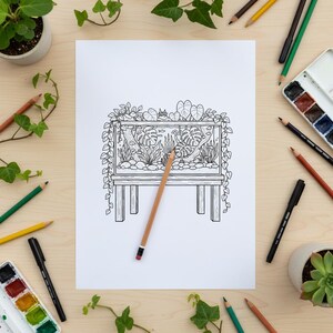 Op de afbeelding: Zwart-witte lijntekening van een aquarium met planten en vissen, rustend op een houten standaard. Een potlood ligt op de tekening. Rondom de tekening liggen kleurpotloden, pennen en aquarelverf.