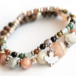 May include: Two beaded bracelets with a variety of colourful stones and silver accents. One bracelet features a silver maple leaf charm. The beads are in shades of brown, green, black, peach, and white. The bracelets are on a white background.