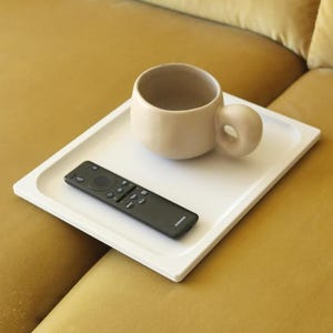 May include: A white rectangular tray holding a beige ceramic mug with a thick rounded handle and a black Samsung remote control. The tray features a smooth surface with slightly raised edges, providing a stable base for the mug and remote.