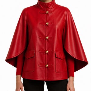 Puede incluir: Chaqueta capa de cuero rojo vibrante con cuello mandarín y tres juegos de cierres de botones dorados. La chaqueta presenta dos bolsillos delanteros y un diseño único tipo capa. Las mangas son largas y la chaqueta es de color rojo liso.