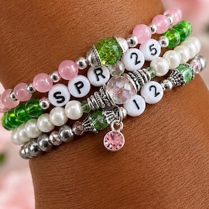 May include: A stack of beaded bracelets in pink, green, white, and silver. One bracelet spells "SPR" with white letter beads. Other bracelets have the numbers "25" and "12". A pink charm hangs from one bracelet.