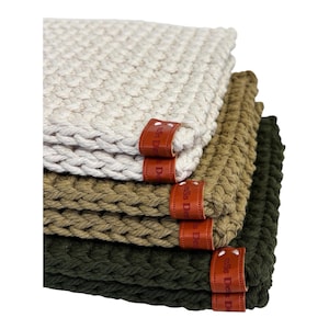 May include: A stack of four woven coasters in varying shades of cream, tan, olive, and dark green. Each coaster features a thick, textured weave and a small, brown leather tag with white text. The coasters are stacked neatly, showcasing the different colors.