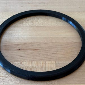 Op de afbeelding: Een zwart, cirkelvormig object met een dik, afgerond profiel. Het object heeft de vorm van een ring, waarschijnlijk gemaakt van plastic of een vergelijkbaar materiaal. De ring ligt op een lichtbruin houten oppervlak.