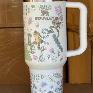 May include: White Stanley travel mug with a handle, featuring a whimsical design of frogs, flowers, and butterflies. The mug has a silver lid and the Stanley logo. The mug is approximately 7 inches tall.