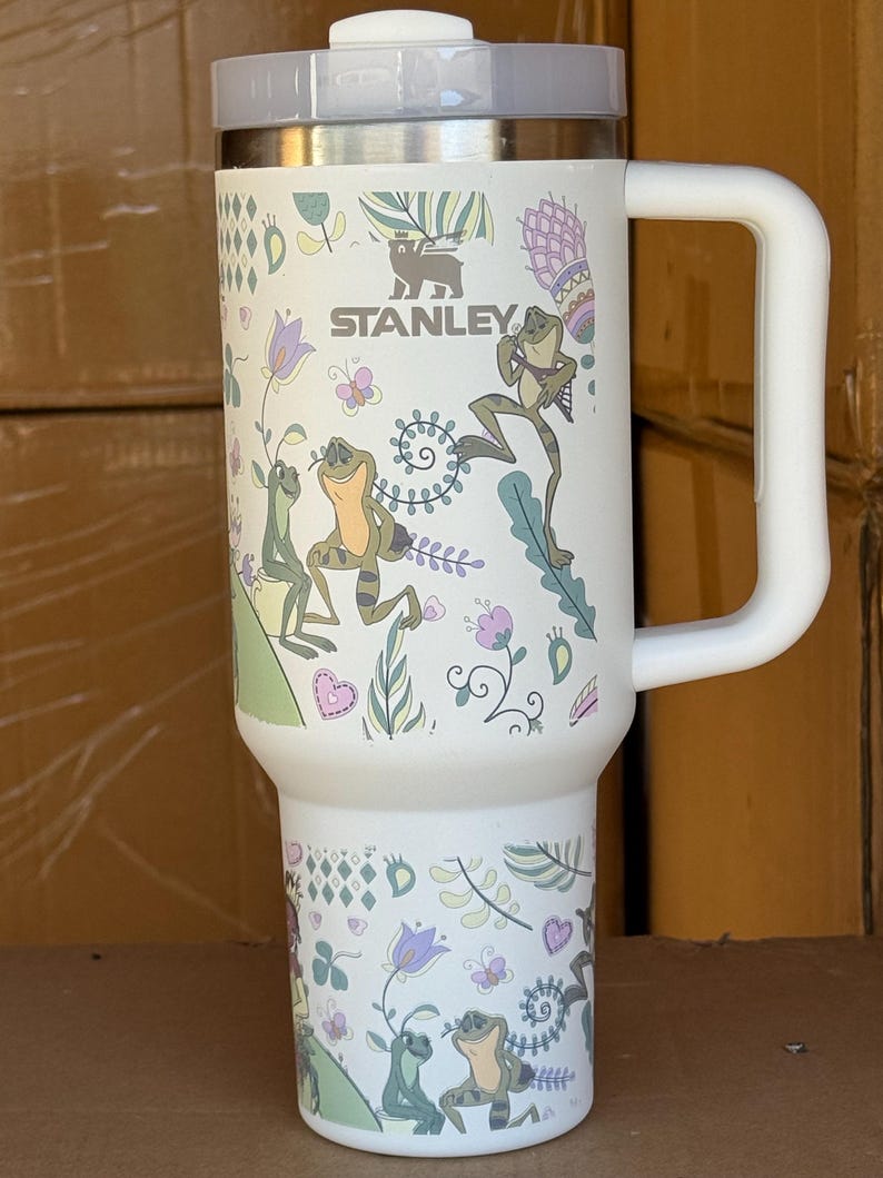 May include: White Stanley travel mug with a handle, featuring a whimsical design of frogs, flowers, and butterflies. The mug has a silver lid and the Stanley logo. The mug is approximately 7 inches tall.