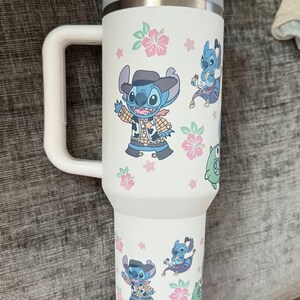 Puede incluir: Vaso t&eacute;rmico blanco con asa y tapa azul claro. Decorado con dibujos animados de Stitch, flores rosas y hojas verdes. El asa es blanca y el borde es de acero inoxidable.
