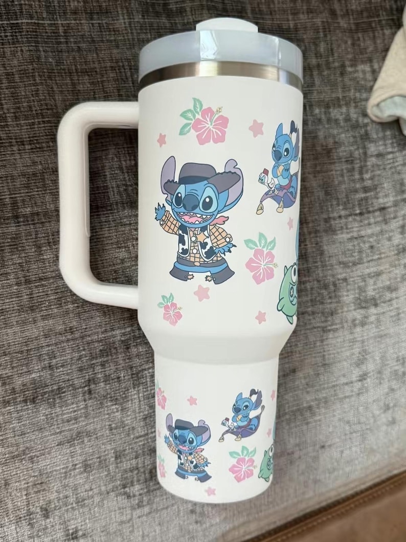 Puede incluir: Vaso t&eacute;rmico blanco con asa y tapa azul claro. Decorado con dibujos animados de Stitch, flores rosas y hojas verdes. El asa es blanca y el borde es de acero inoxidable.
