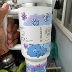 May include: White Stanley Quencher tumbler with a handle, featuring a floral and Stitch design. The tumbler has a white lid and a stainless steel band. The design includes blue and purple flowers and a cartoon character. It holds 40 oz.