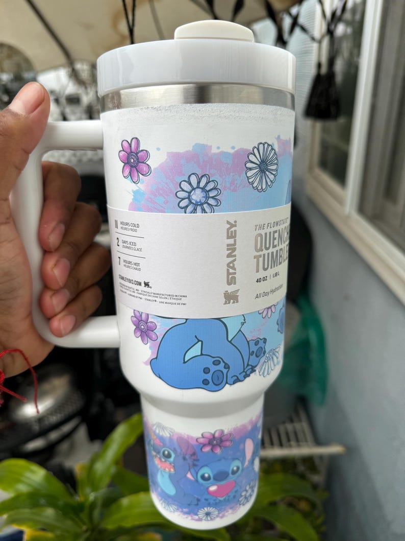 May include: White Stanley Quencher tumbler with a handle, featuring a floral and Stitch design. The tumbler has a white lid and a stainless steel band. The design includes blue and purple flowers and a cartoon character. It holds 40 oz.