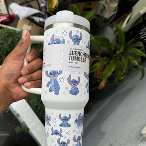 May include: White Stanley Quencher H2.0 Flowstate tumbler featuring a repeating pattern of the character Stitch from Lilo & Stitch. The tumbler has a white handle and lid. The text "QUENCHER TUMBLER" and "40 OZ | 1.18 L" is visible.