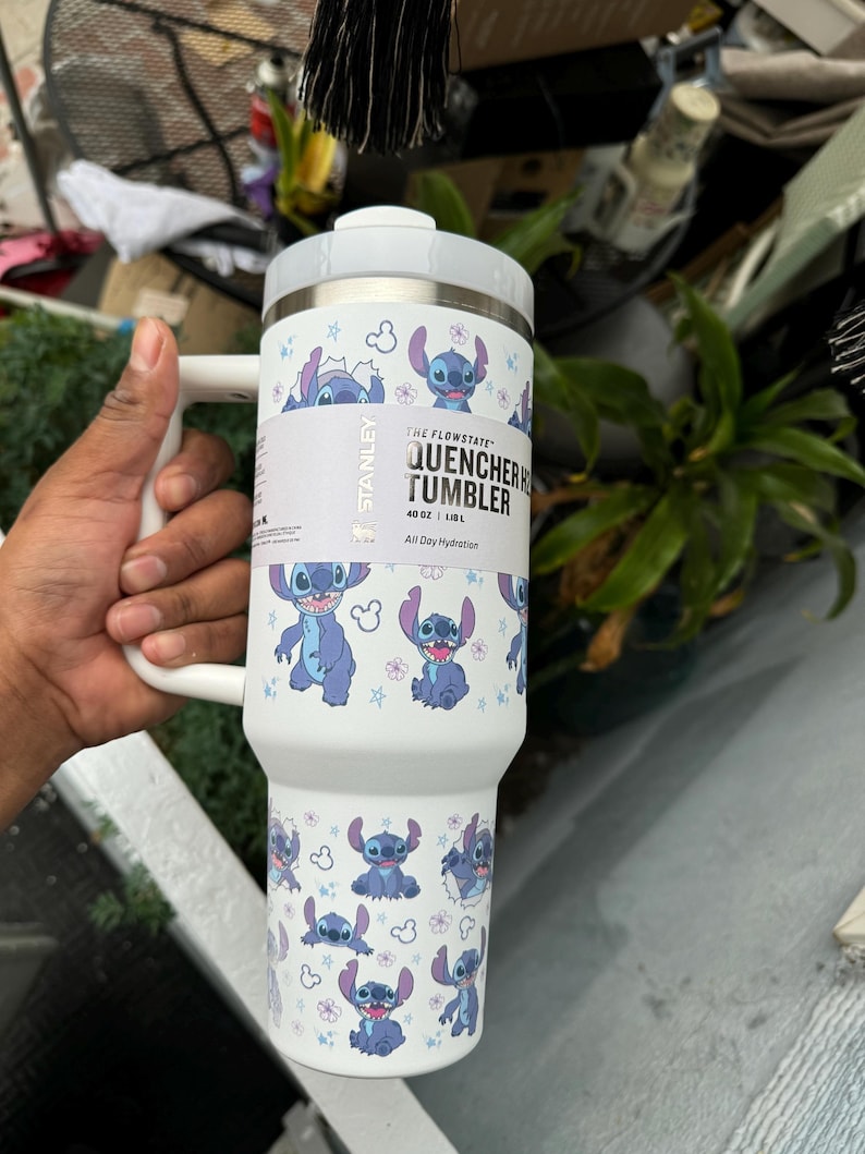 May include: White Stanley Quencher H2.0 Flowstate tumbler featuring a repeating pattern of the character Stitch from Lilo & Stitch. The tumbler has a white handle and lid. The text "QUENCHER TUMBLER" and "40 OZ | 1.18 L" is visible.
