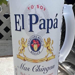 May include: Frosted white beer mug with the text "El Papá" in blue, featuring a crest with lions and the words "Cerveceria Modelo Mexico 1925". The mug also has the text "Mas Chingon".