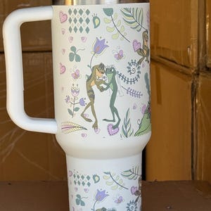 May include: White insulated tumbler with a handle and a lid. The tumbler is decorated with a whimsical design featuring frogs, flowers, and hearts. The design includes green, purple, and pink accents.