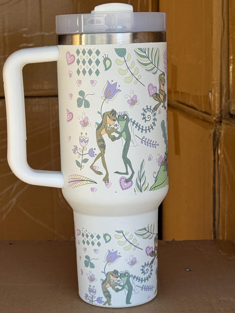 May include: White insulated tumbler with a handle and a lid. The tumbler is decorated with a whimsical design featuring frogs, flowers, and hearts. The design includes green, purple, and pink accents.