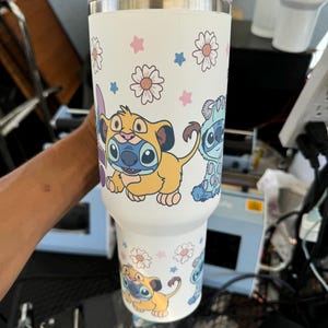 Puede incluir: Vaso t&eacute;rmico blanco con personajes de dibujos animados y dise&ntilde;os florales. El vaso tiene una tapa blanca y un borde de acero inoxidable. El dise&ntilde;o incluye un personaje amarillo y azul, junto con estrellas y flores rosas y azules.