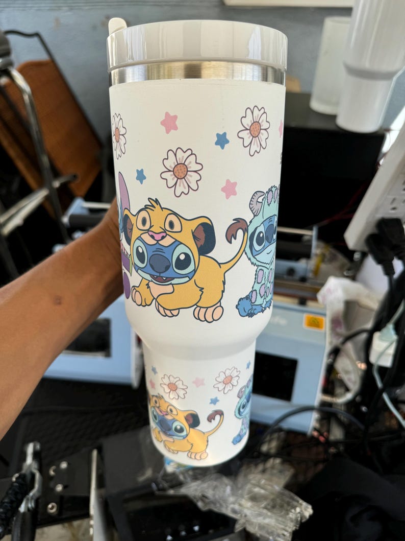 Puede incluir: Vaso t&eacute;rmico blanco con personajes de dibujos animados y dise&ntilde;os florales. El vaso tiene una tapa blanca y un borde de acero inoxidable. El dise&ntilde;o incluye un personaje amarillo y azul, junto con estrellas y flores rosas y azules.
