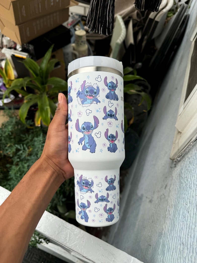 May include: White insulated tumbler featuring a repeating pattern of the character Stitch from the animated film Lilo & Stitch. The design includes Stitch in various poses, surrounded by flowers and stars. The tumbler has a white lid and a stainless steel body.