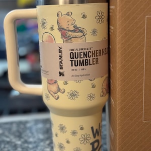 Puede incluir: Un vaso Stanley Quencher H2.0 amarillo con asa, con ilustraciones de Winnie the Pooh, abejas y flores. El vaso tiene una capacidad de 1,18 L e incluye el texto "THE FLOWSTATE QUENCHER H2.0 TUMBLER".