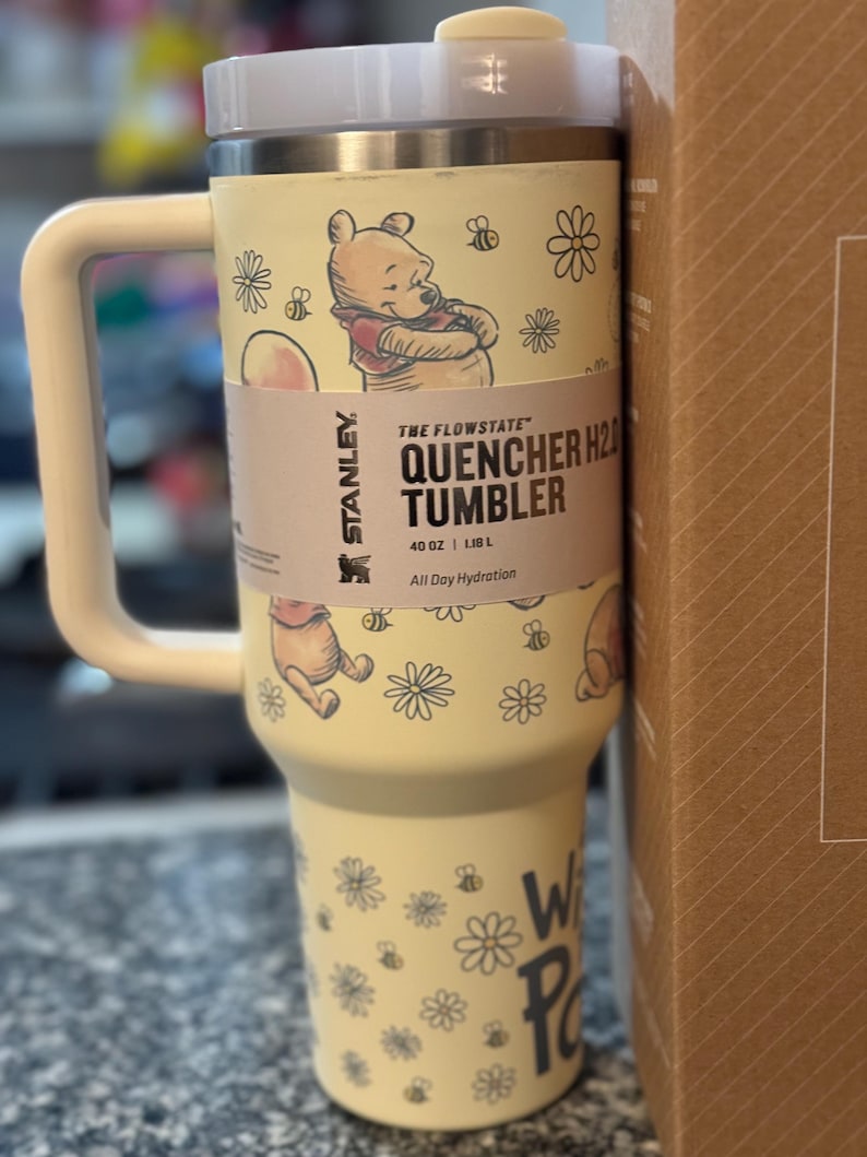 Puede incluir: Un vaso Stanley Quencher H2.0 amarillo con asa, con ilustraciones de Winnie the Pooh, abejas y flores. El vaso tiene una capacidad de 1,18 L e incluye el texto "THE FLOWSTATE QUENCHER H2.0 TUMBLER".