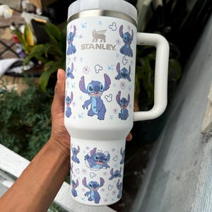May include: White Stanley tumbler featuring a repeating pattern of the character Stitch from Lilo & Stitch. The tumbler has a white handle and lid, with the Stanley logo visible. The design includes blue Stitch illustrations and floral accents.