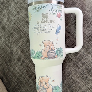May include: White Stanley travel mug featuring Winnie the Pooh illustrations. The mug has a light blue lid and handle, with the text "Sometimes the smallest things take up the most room in your heart." The mug has a capacity of approximately 30 ounces.