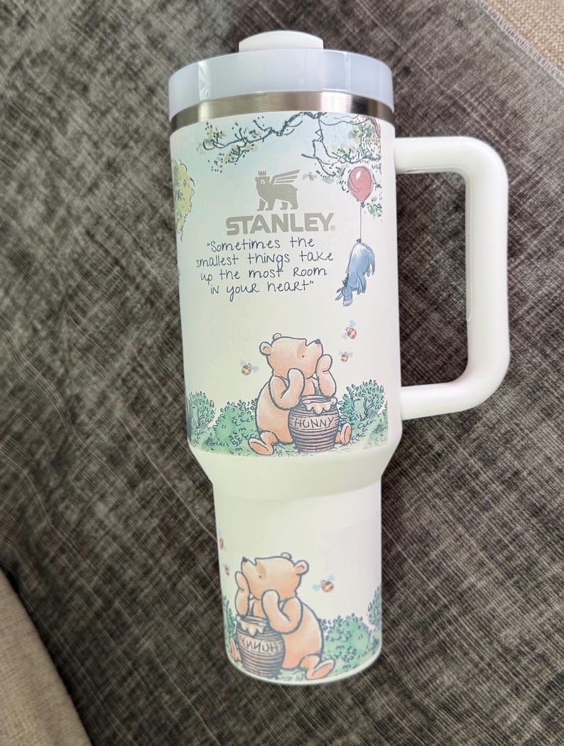 May include: White Stanley travel mug featuring Winnie the Pooh illustrations. The mug has a light blue lid and handle, with the text "Sometimes the smallest things take up the most room in your heart." The mug has a capacity of approximately 30 ounces.