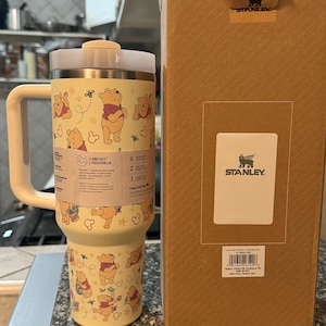May include: A yellow Stanley Flowstate tumbler with a Winnie the Pooh design, featuring the character and bees. The tumbler has a white handle and lid, and is next to a brown Stanley box with the logo.
