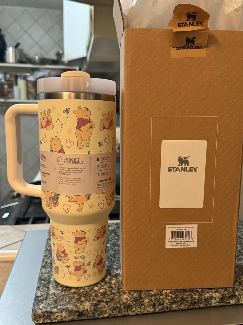 May include: A yellow Stanley Flowstate tumbler with a Winnie the Pooh design, featuring the character and bees. The tumbler has a white handle and lid, and is next to a brown Stanley box with the logo.