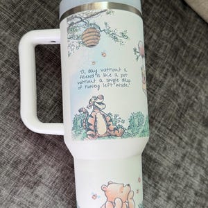 May include: White insulated tumbler with a light blue lid and handle. The tumbler features illustrations of Winnie the Pooh characters, a beehive, and the quote "A day without a friend is like a pot without a single drop of honey left inside."