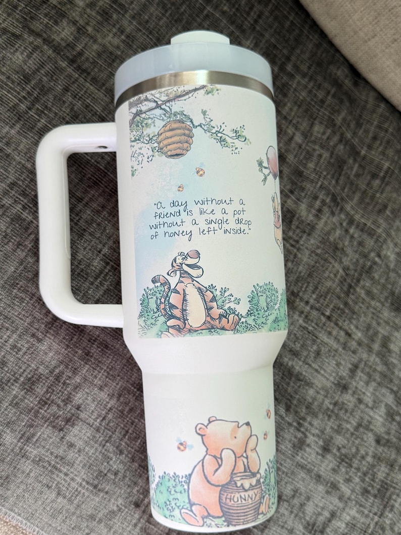 May include: White insulated tumbler with a light blue lid and handle. The tumbler features illustrations of Winnie the Pooh characters, a beehive, and the quote "A day without a friend is like a pot without a single drop of honey left inside."
