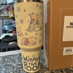 Puede incluir: Un vaso Stanley amarillo con ilustraciones de Winnie the Pooh. El vaso tiene una tapa blanca y un cuerpo amarillo claro con dibujos de Winnie the Pooh, abejas y flores. Las palabras "Winnie the Pooh" est&aacute;n impresas en la parte inferior.