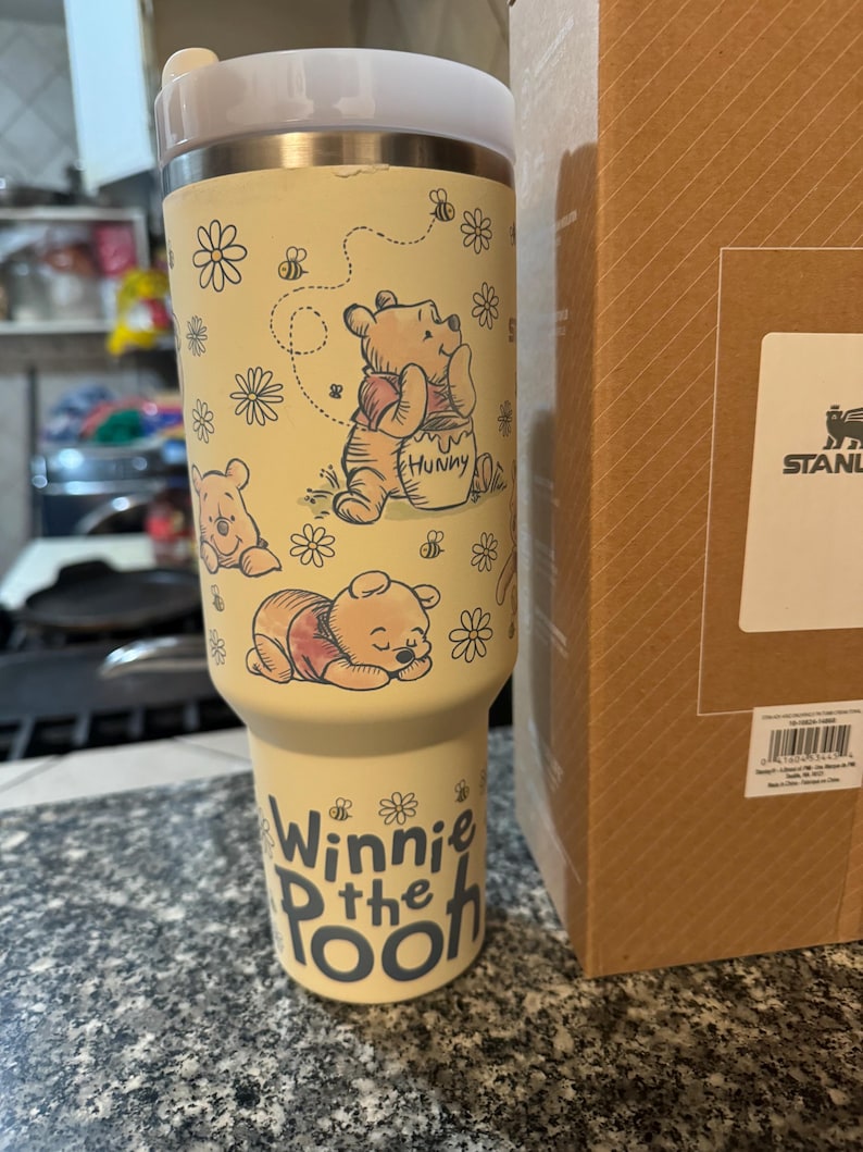 Puede incluir: Un vaso Stanley amarillo con ilustraciones de Winnie the Pooh. El vaso tiene una tapa blanca y un cuerpo amarillo claro con dibujos de Winnie the Pooh, abejas y flores. Las palabras "Winnie the Pooh" est&aacute;n impresas en la parte inferior.