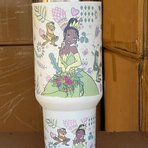 May include: A white insulated tumbler featuring a cartoon illustration of a princess in a green dress, holding flowers, and a frog. The design includes hearts, flowers, and decorative elements. The tumbler has a gray lid.