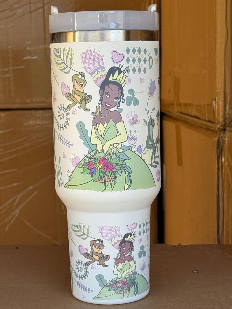 May include: A white insulated tumbler featuring a cartoon illustration of a princess in a green dress, holding flowers, and a frog. The design includes hearts, flowers, and decorative elements. The tumbler has a gray lid.