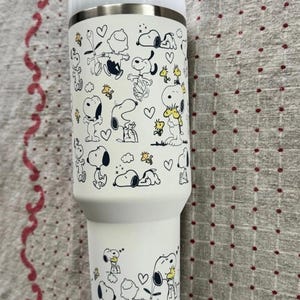 Puede incluir: Un vaso t&eacute;rmico blanco con un patr&oacute;n repetido de los personajes de Snoopy y Woodstock en negro y amarillo. El vaso tiene un borde de acero inoxidable y una tapa blanca. El dise&ntilde;o incluye corazones y otros gr&aacute;ficos peque&ntilde;os.
